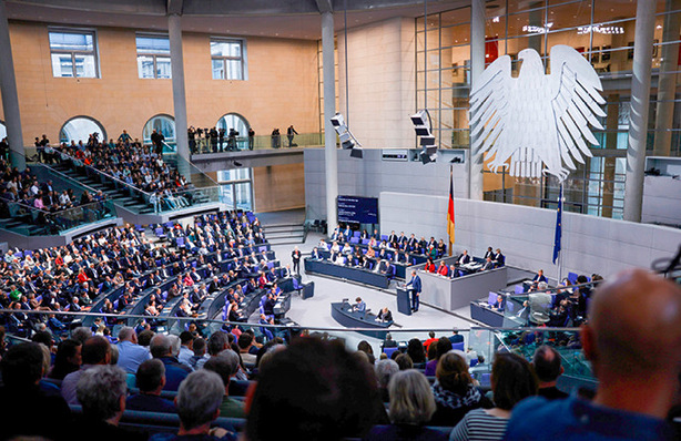 Sitzung des Deutschen Bundestages in einem großen, modernen Saal mit sitzenden Menschen; an der Wand ist ein riesiges Adleremblem zu sehen. - © Foto: Deutscher Bundestag/Tobias Koch