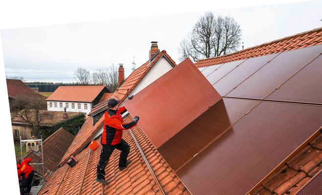 Arbeiter in Rot installiert Solarmodule auf einem roten Ziegeldach in einer ländlichen Gegend mit Bäumen und Häusern im Hintergrund. - © Foto: BSH GmbH & Co. KG