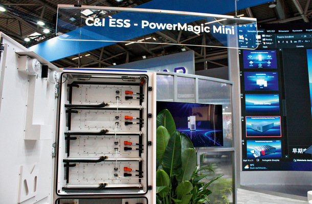 Offene Vitrine mit PowerMagic Mini-Energiespeichern auf einer Technologiemesse, mit digitalen Anzeigen im Hintergrund. - © Foto: Heiko Schwarzburger