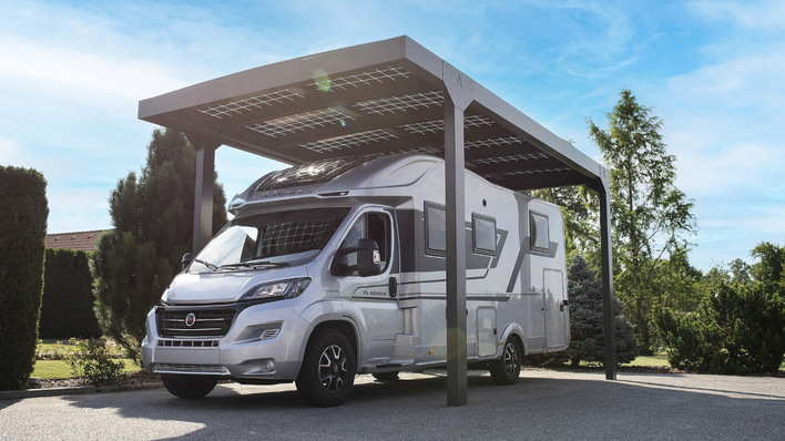 Ein silbernes Wohnmobil, geparkt unter einem modernen Carport mit Sonnenkollektoren auf dem Dach, umgeben von Bäumen. - © Alukov